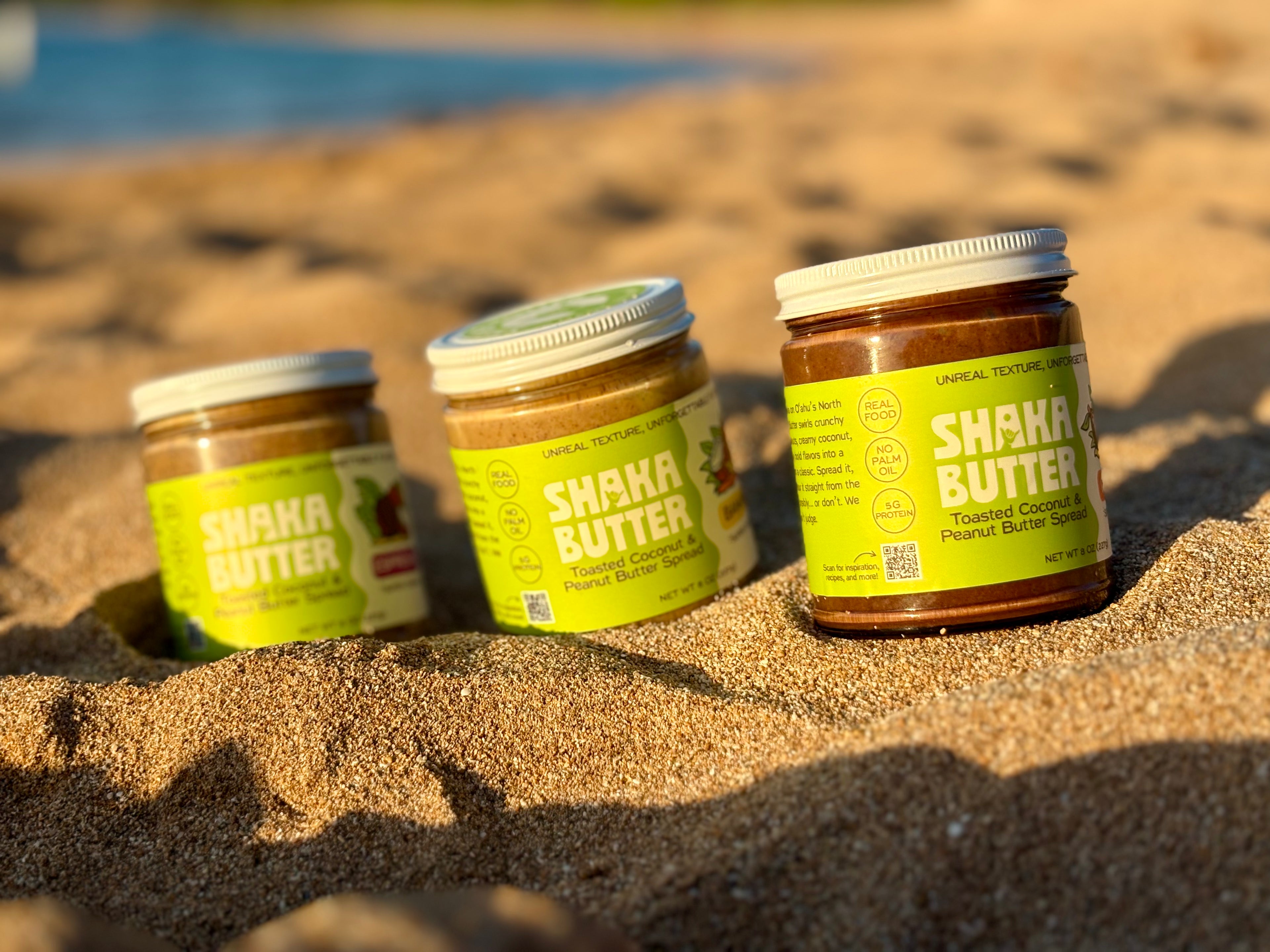 Jars of Shaka Butter on a sandy surface with a blurred natural background